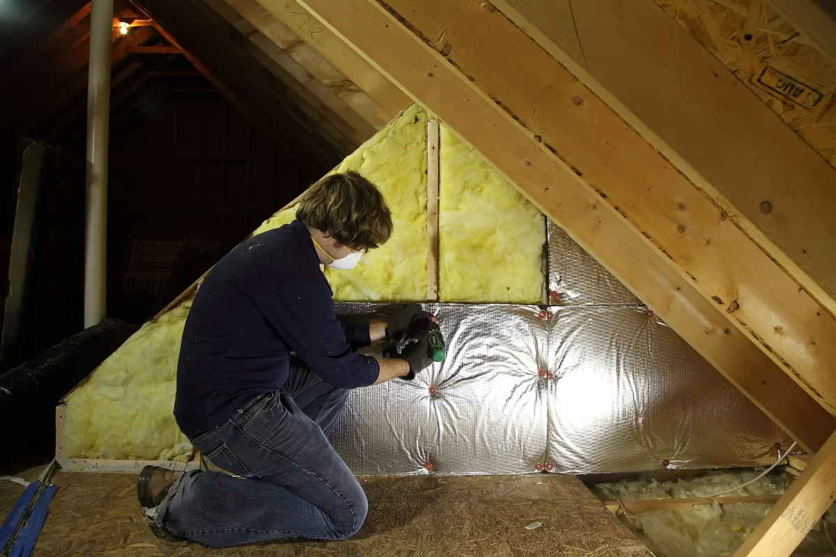 A man in a dust mask fixing foil-backed insulation boards between the rafters of a loft